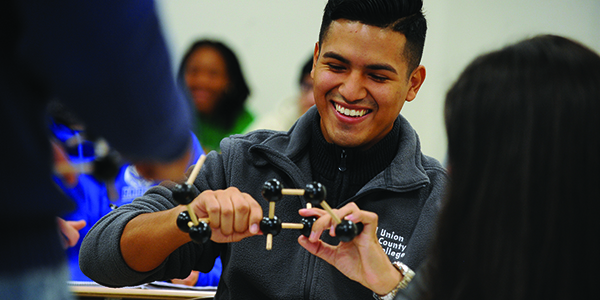 A student smiling while holding an object in a classroom.
