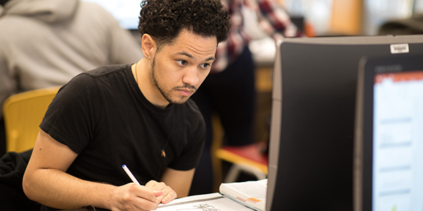 A student writing on paper while looking at a computer screen.