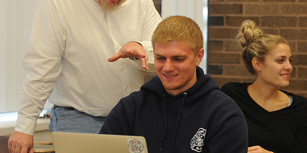 A professor talking to a student who is working on a laptop.